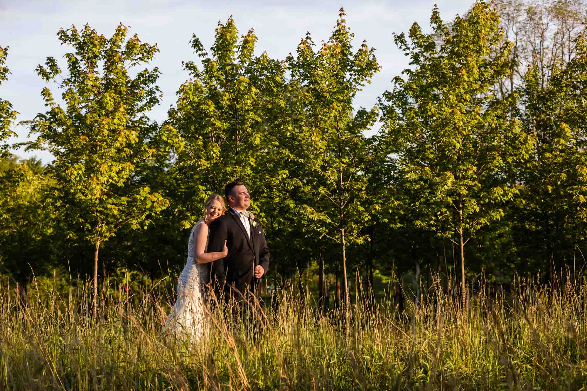 bride and groom snuggle together in front of a row of trees at sunset